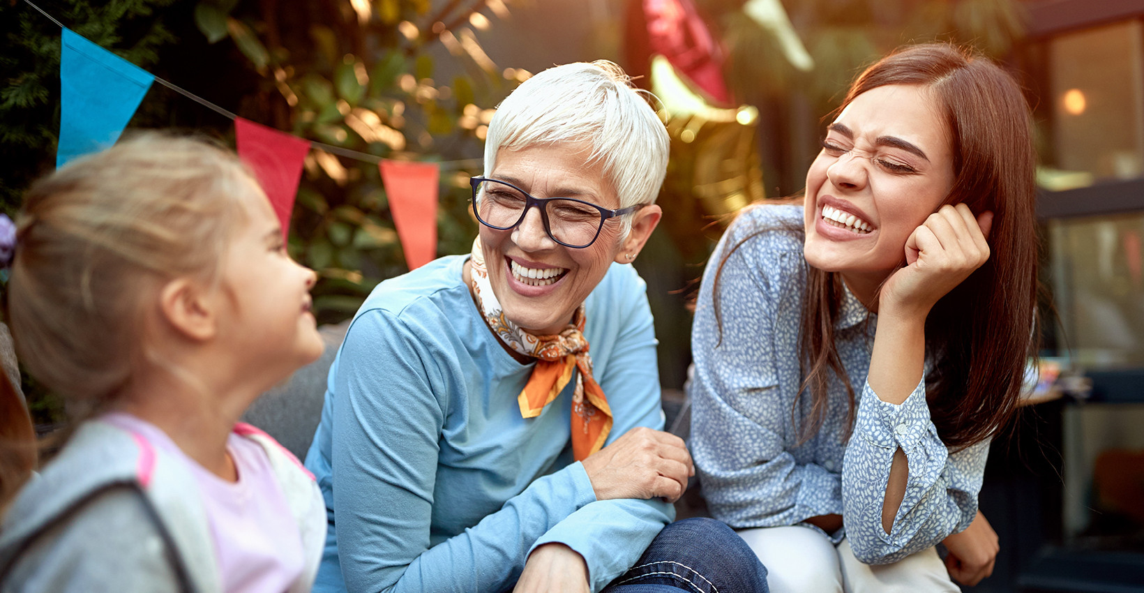 An elderly lady, her daughter, and her granddaughter laughing in a backyard