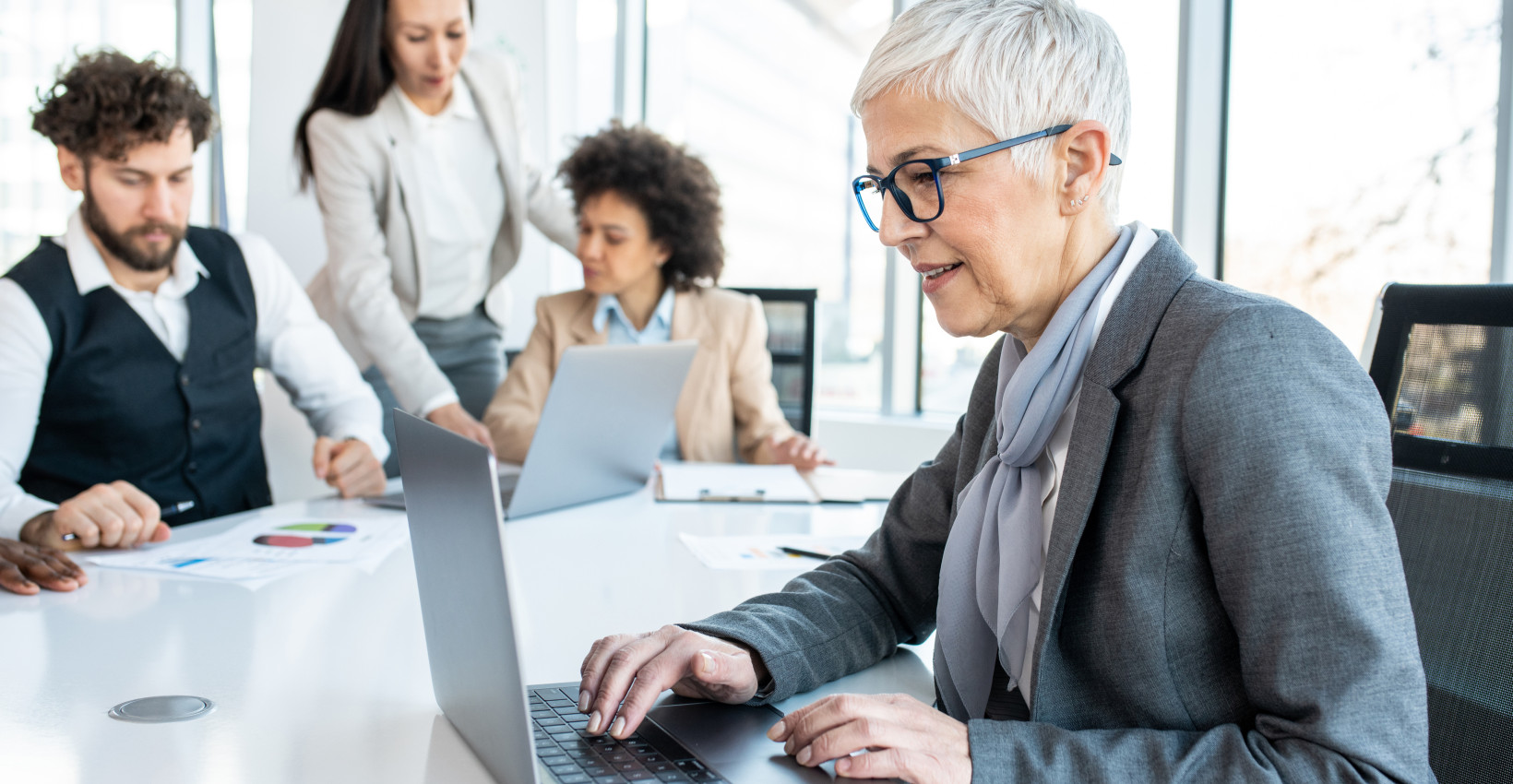 a woman sits at a computer at a conference table with other coworkers