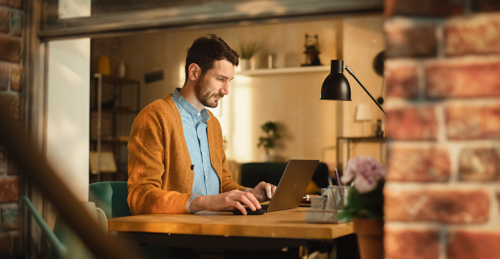 a man sits at his laptop on a wooden desk with a brick wall