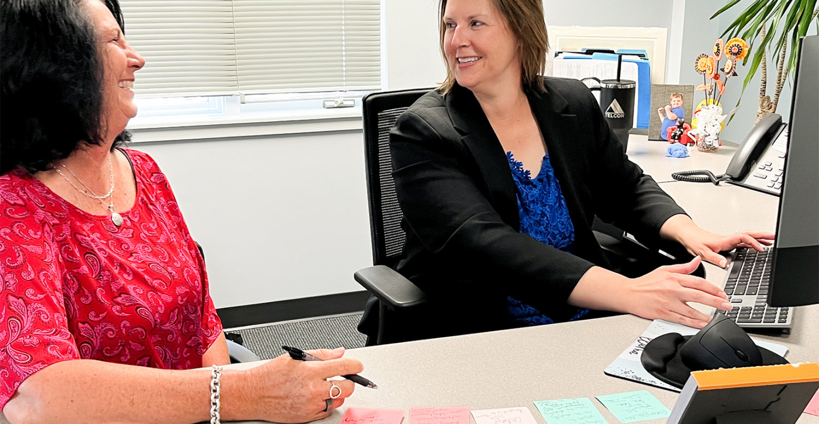 two women sit at a desk. one wears a red shirt the other wears a black shirt