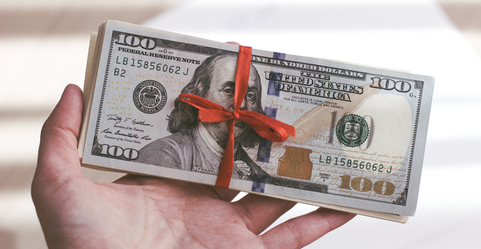 a hand holds a hundred dollar bill wrapped in red ribbon