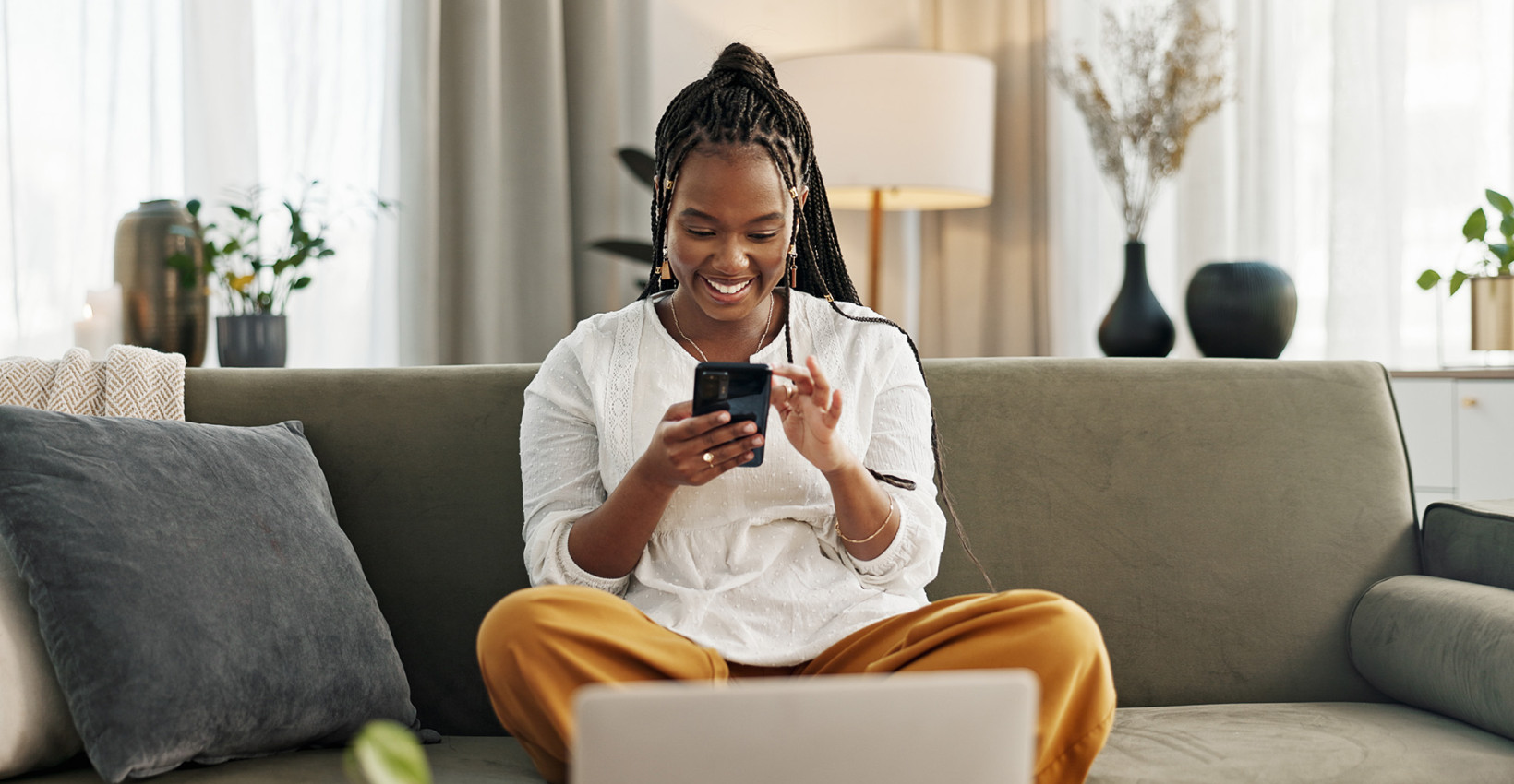 A woman sits on a her couch with her laptop in front of her while holding her phone
