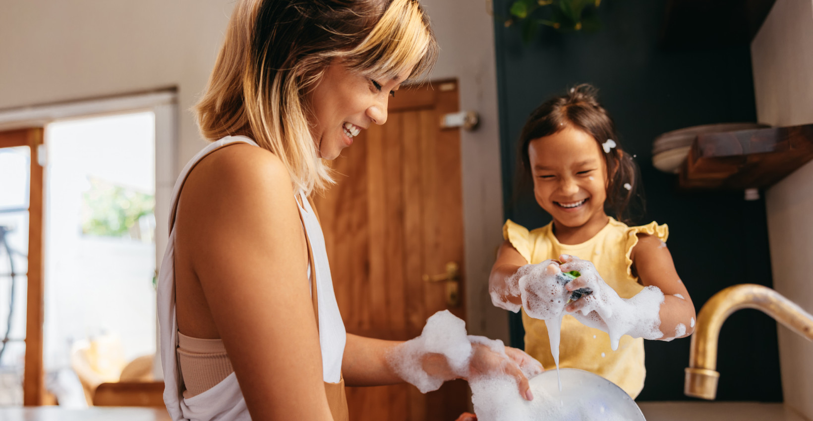 a mom stands at the sink with her daughter who is playing in soap bubbles