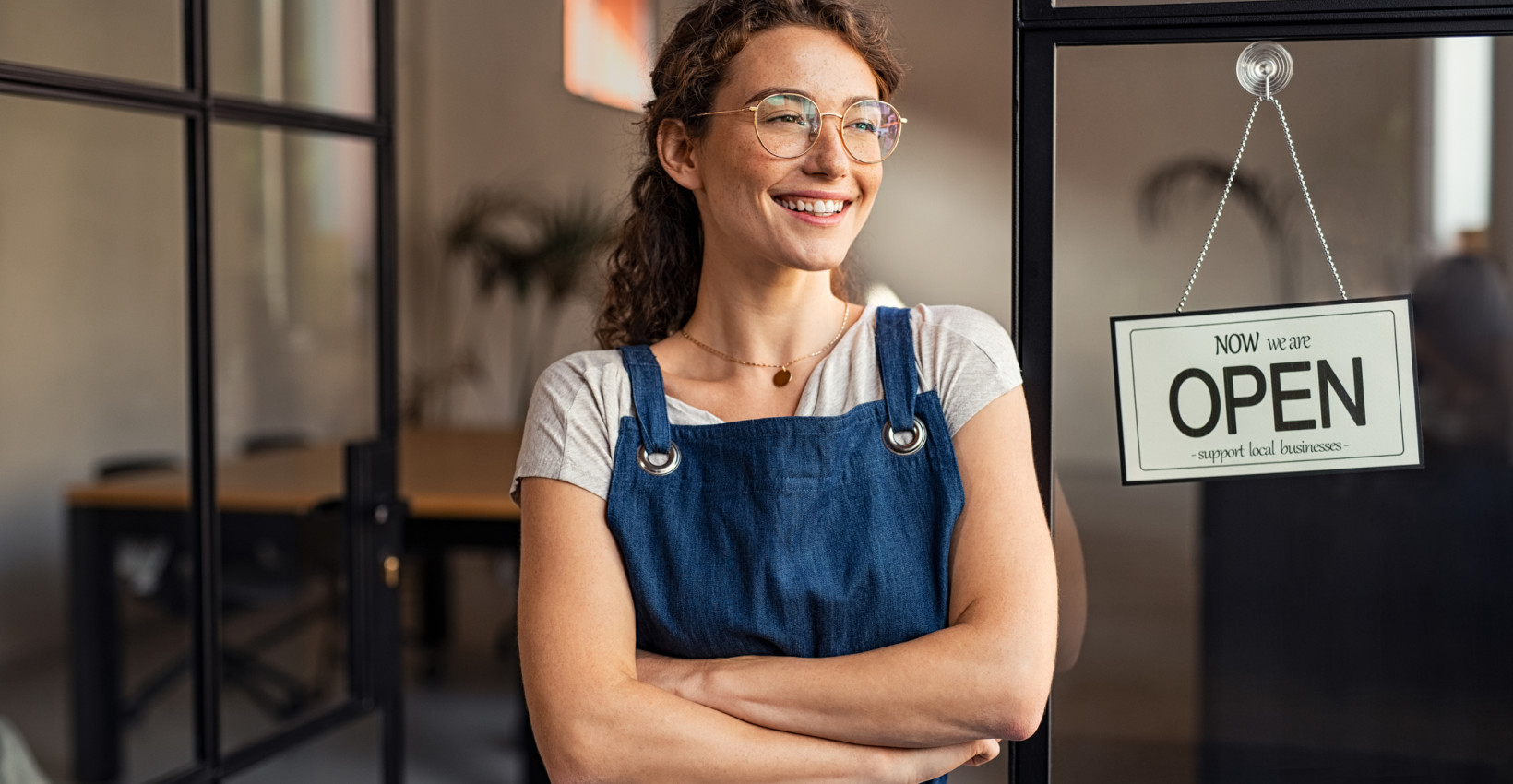 a woman with glasses wearing a blue apron stands in a door way next to a sign that says open