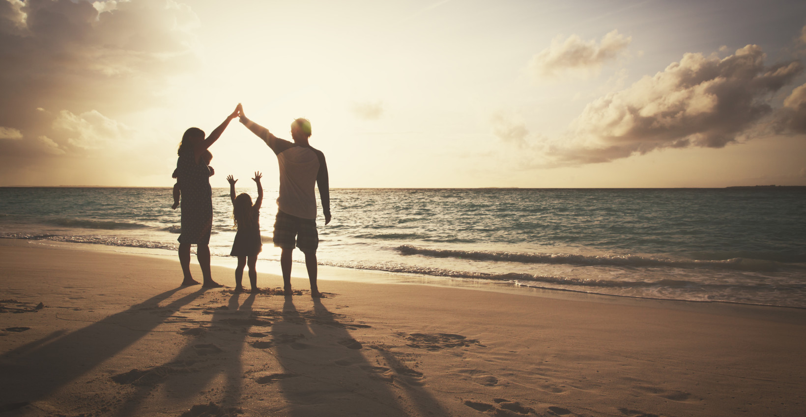 three people stand on a beach next to an ocean in shadow holding their arms above their heads