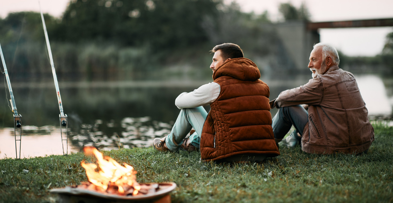 a dad and son sit together on the grass next to a fire