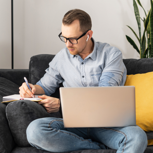 A man sitting on the couch with a laptop on his lap while he writes something in a notebook