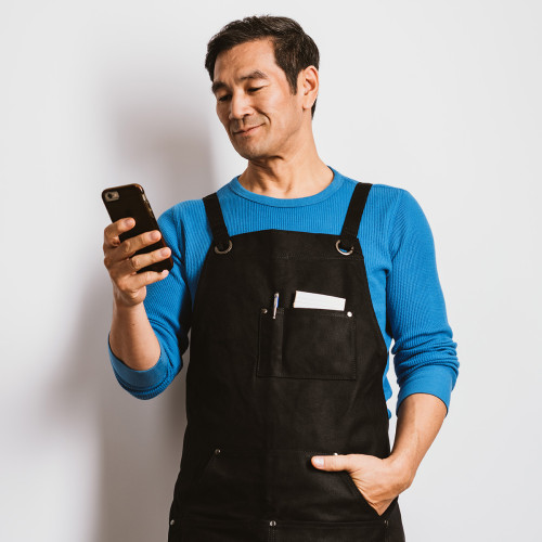 a man in a black apron stands holding his phone smiling