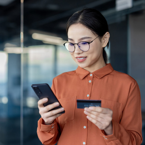 A woman wearing an orange shirt smiles as she looks down at her phone and debit card