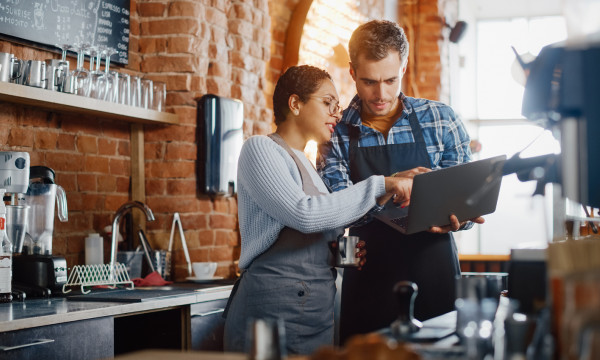 A man an dwoman looking at a laptop behind the counter of a coffee shop