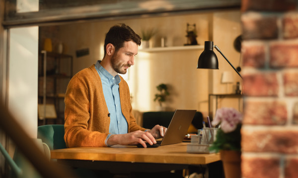 a man sits at his laptop on a wooden desk with a brick wall