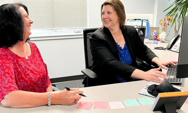 two women sit at a desk. one wears a red shirt the other wears a black shirt