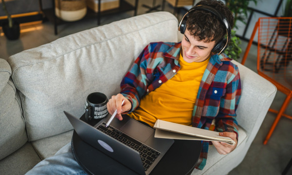 a teen lays on the couch with his laptop on his lap and holds a notebook while wearing headphones