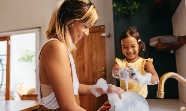 a mom stands at the sink with her daughter who is playing in soap bubbles
