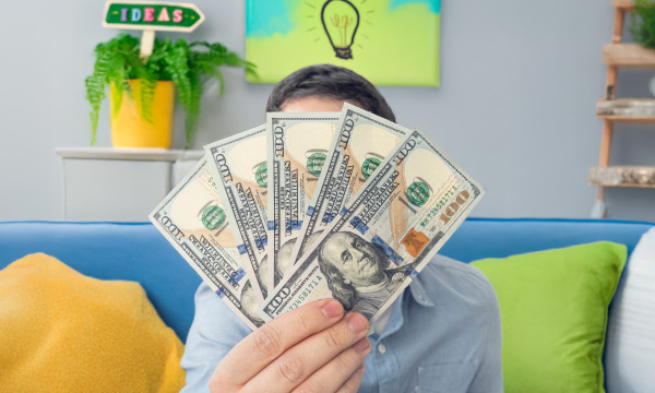 a person wearing a blue shirt holds up five 100 dollar bills while sitting on a couch
