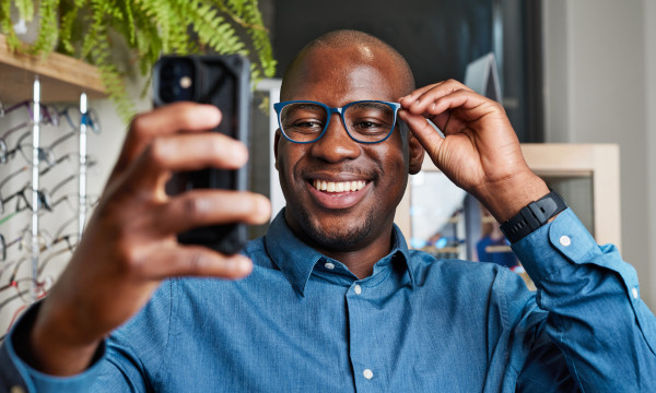 a black man wearing a blue button up shirt smiles while taking a selfie on his phone
