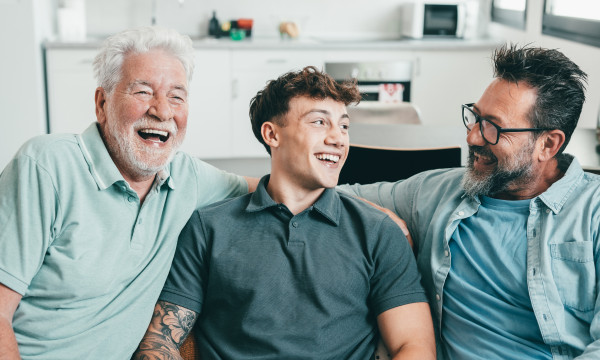 a young, middle-aged, and older man sit together on a couch laughing