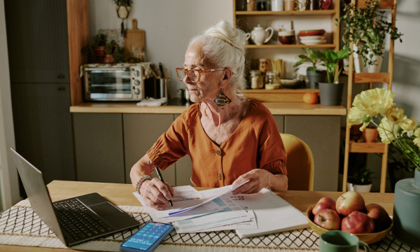an elderly woman sits at a desk with paperwork and a laptop