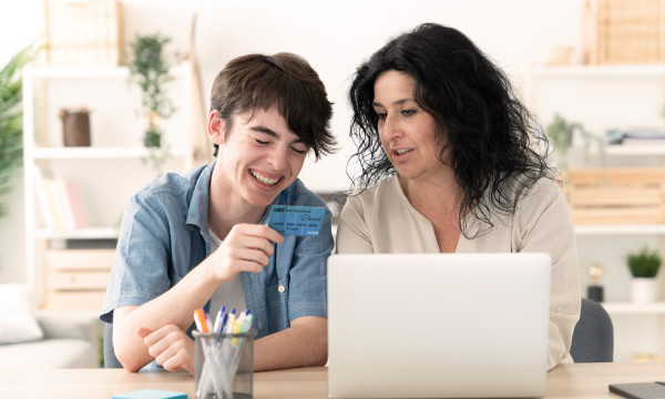 a mother and son sit at a laptop together laughing. the son holds a credit card