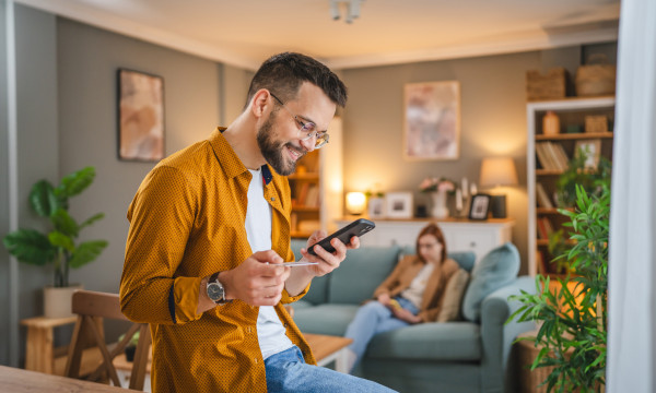 a man leans on a table holding his phone and debit card while a woman sits on the couch behind him