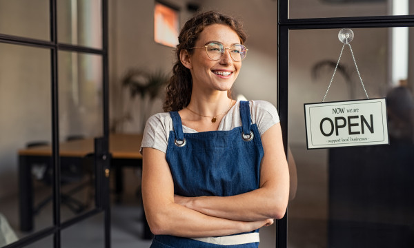 a woman with glasses wearing a blue apron stands in a door way next to a sign that says open
